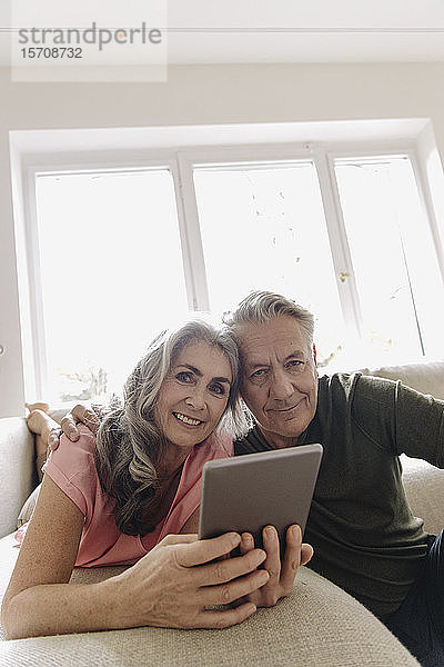 Porträt eines glücklichen älteren Paares,  das sich zu Hause auf der Couch mit Tablette entspannt