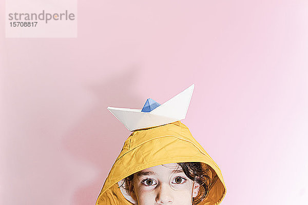 Kleines Mädchen,  das ein Ölzeug trägt und ein Papierboot auf dem Kopf trägt