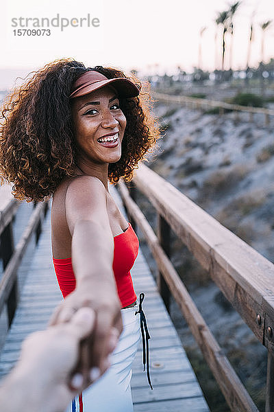 Lächelnde junge Frau mit brauner Mütze führt ihre beste Freundin an den Strand