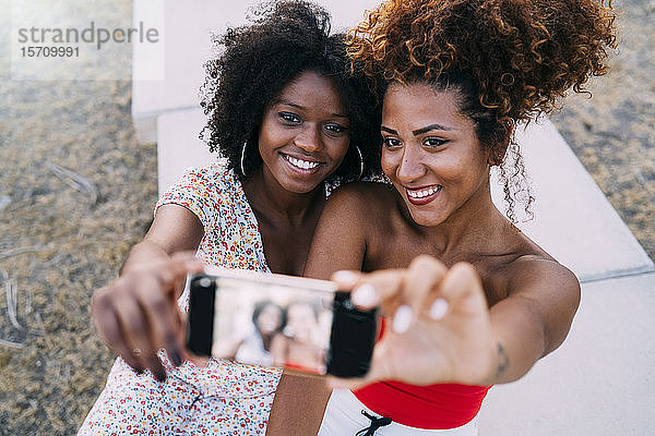 Junge Frauen,  die lächeln und sich mit ihrem Smartphone in einem Park ein Selfie machen,  Fokus auf den Hintergrund