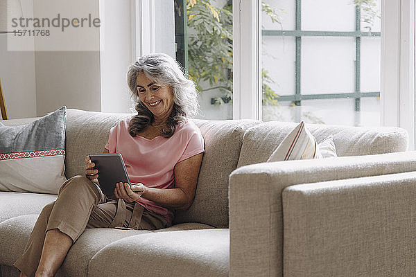 Lächelnde reife Frau mit Tablette zu Hause auf dem Sofa sitzend