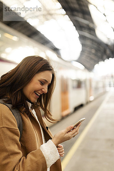 Glückliche junge Frau benutzt Mobiltelefon am Bahnhof