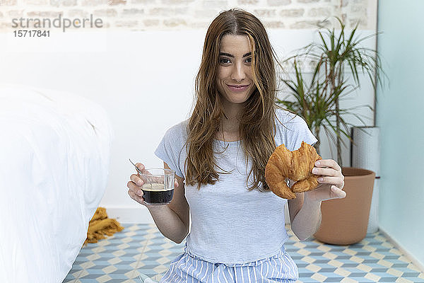 Junge Frau sitzt auf dem Boden und hält eine Tasse Kaffee und ein Croissant