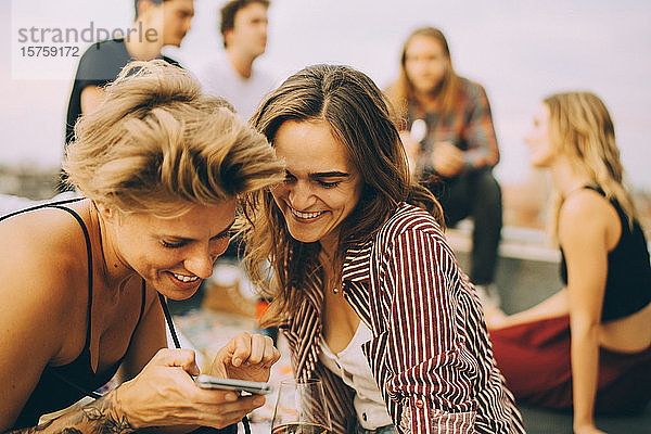 Lächelnde Frau zeigt einem Freund ihr Handy,  während sich die Leute während der Party auf der Gebäudeterrasse vergnügen