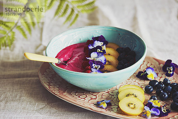 Hochwinkel-Nahaufnahme einer Schale mit rosa Fruchtpüree mit Heidelbeeren und Scheiben von gelben Kiwis,  dekoriert mit Stiefmütterchenblättern.