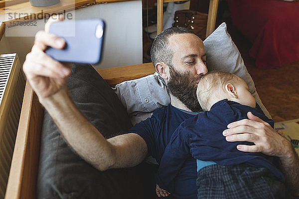 Mann,  der sich mit seinem niedlichen schlafenden Sohn auf dem Sofa ein Selfie nimmt