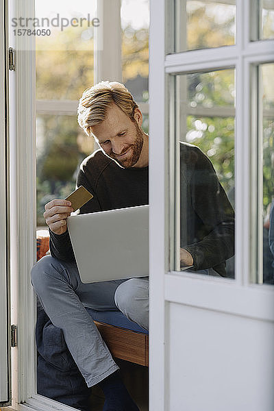 Lächelnder Mann mit Laptop und Kreditkarte im Wintergarten zu Hause
