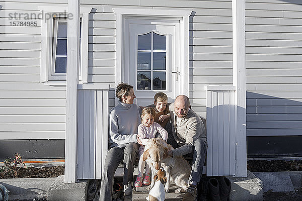 Glückliche Familie mit zwei Kindern und zwei Hunden,  die vor ihrem Haus sitzen