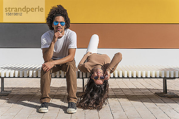 Paar mit Sonnenbrille,  entspannt auf der Bank,  ein Partner auf dem Rücken liegend mit herabhängendem Kopf