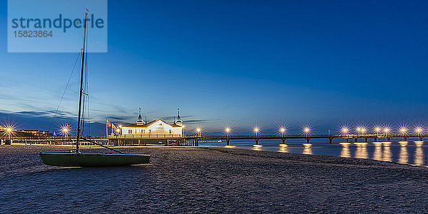 Deutschland,  Mecklenburg-Vorpommern,  Heringsdorf,  Segelboot in der Abenddämmerung am Sandstrand mit beleuchteter Mole im Hintergrund