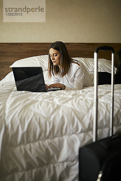 Geschäftsfrau,  die im Hotelzimmer auf dem Bett liegt und einen Laptop benutzt