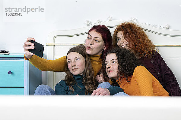 Mutter und ihre drei Töchter sitzen zusammen im Bett und nehmen Selfie mit Smartphone