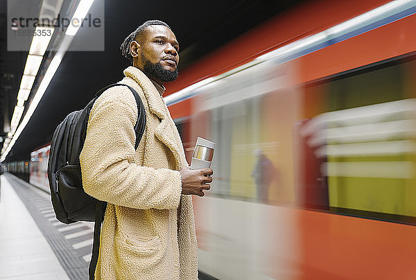 Stilvoller Mann mit wiederverwendbarer Tasse und Kopfhörern in einer U-Bahn-Station