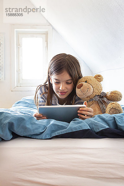 Porträt eines lächelnden Mädchens mit Teddybär auf dem Bett liegend mit digitalem Tablett