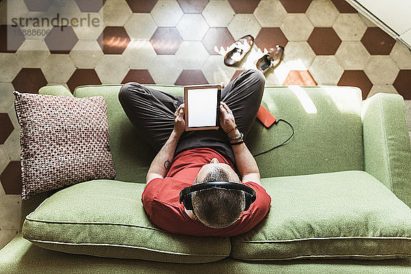 Reifer Mann sitzt im Wohnzimmer auf dem Sofa und benutzt ein Tablett,  hört Musik