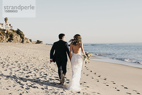 Rückansicht des am Strand laufenden glücklichen Brautpaares