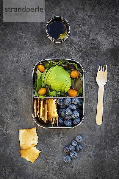 Schale mit Salatdressing und Brotdose mit Avocadoscheiben,  gelben Tomaten,  Crackern,  Blaubeeren und grünem Salat