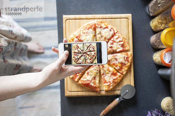 Ausschnittansicht einer Frau,  die eine selbstgemachte Pizza mit einem Smartphone fotografiert