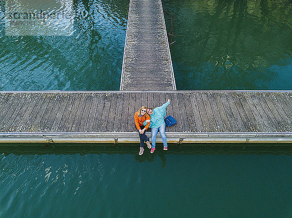 Zwei glückliche Freunde sitzen Seite an Seite auf dem Bootssteg,  Valdemurio-Stausee,  Asturien,  Spanien