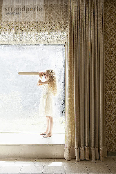 Mädchen steht auf der Fensterbank und schaut durch ein Pappteleskop