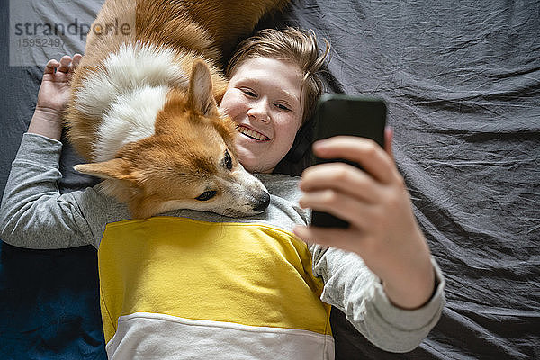 Porträt eines Jungen,  der mit seinem Hund auf dem Bett liegt und mit seinem Smartphone Selfie nimmt