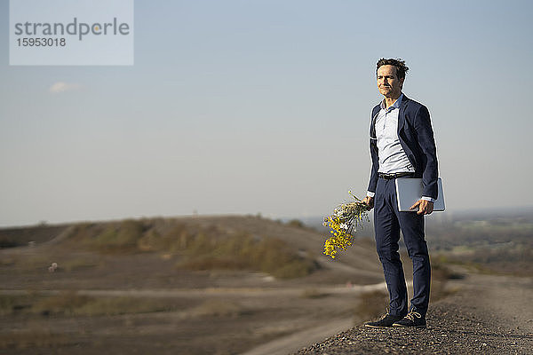 Ein reifer Geschäftsmann hält einen Blumenstrauss und einen Laptop auf einer stillgelegten Minenhalde