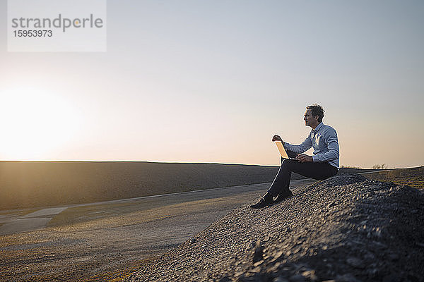 Reifer Geschäftsmann benutzt Laptop auf einer stillgelegten Minenhalde bei Sonnenuntergang