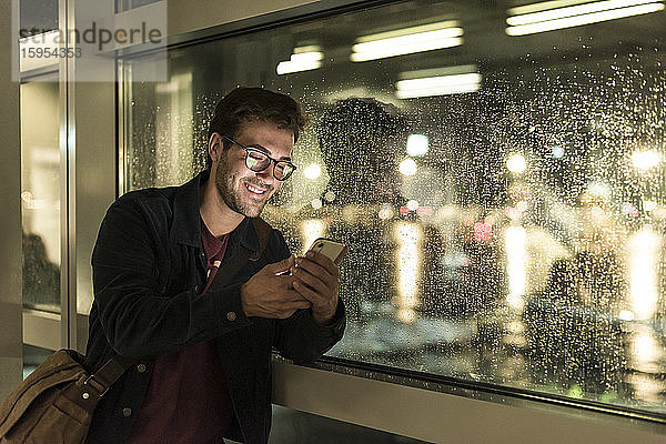 Lächelnder junger Mann benutzt Smartphone bei regnerischem Fenster in der Nacht