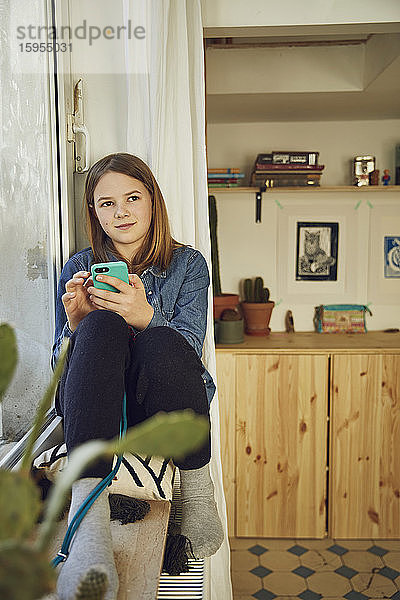 Mädchen sitzt zu Hause am Fenster und hält ein Smartphone