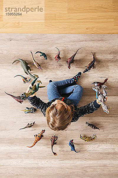 Kleines Mädchen sitzt auf dem Boden und spielt mit Spielzeug-Dinosauriern um sich herum,  Draufsicht