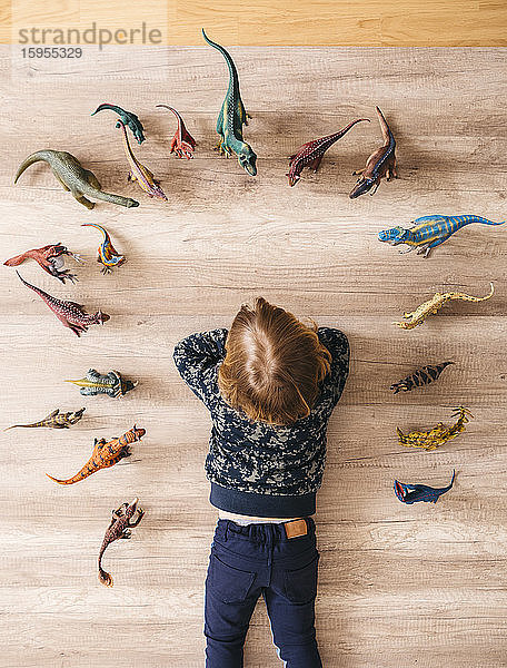 Kleines Mädchen liegt auf dem Boden und spielt mit Spielzeug-Dinosauriern um sich herum,  Draufsicht