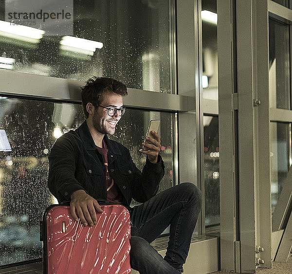 Lächelnder junger Mann mit Koffer sitzt am verregneten Fenster und benutzt ein Smartphone
