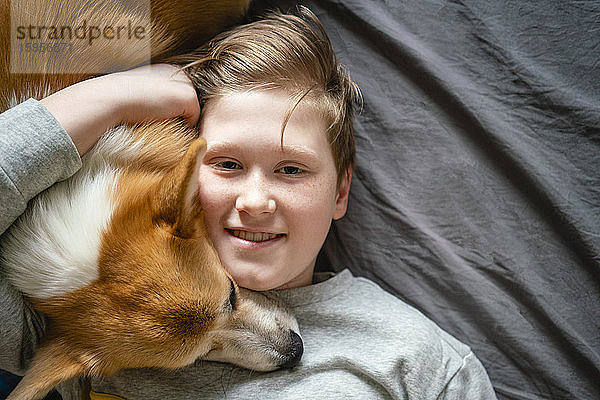 Porträt eines lächelnden Jungen,  der auf dem Bett liegt und seinen Hund kuschelt