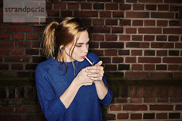 Junge Frau zündet nachts eine Zigarette gegen eine Ziegelmauer