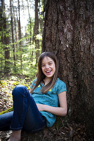 Porträt eines lachenden Mädchens,  das sich im Wald an einen Baumstamm lehnt