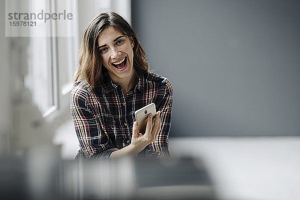Porträt einer lachenden jungen Frau mit Smartphone in einem Loft