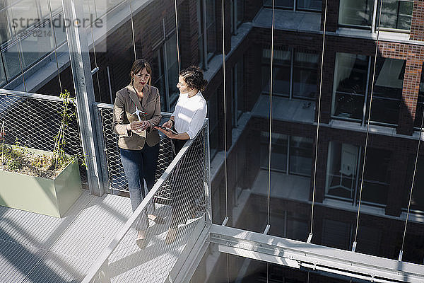 Zwei Geschäftsfrauen mit Tablet und Windradmodell bei einem Treffen auf dem Balkon eines Bürogebäudes