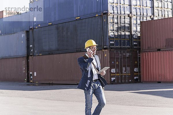Geschäftsmann am Telefon mit digitalem Tablet vor Frachtcontainern gehend