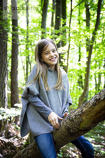 Porträt eines lächelnden niedlichen Mädchens,  das auf einem Baumstamm im Wald sitzt