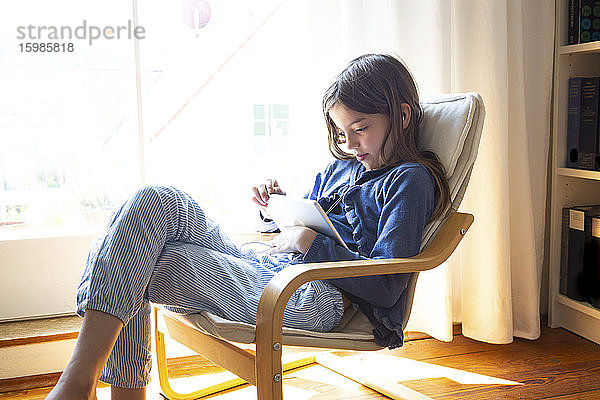 Junges Mädchen sieht sich ein Online-Video auf einem digitalen Tablet an,  während sie zu Hause am Fenster sitzt
