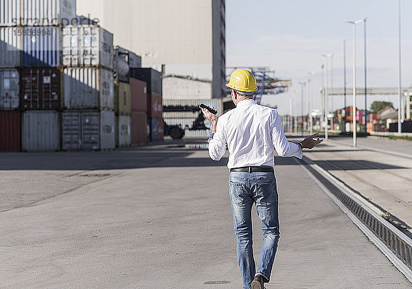 Rückenansicht eines Geschäftsmannes mit Schutzhelm,  der auf einem Industriegelände ein Mobiltelefon benutzt