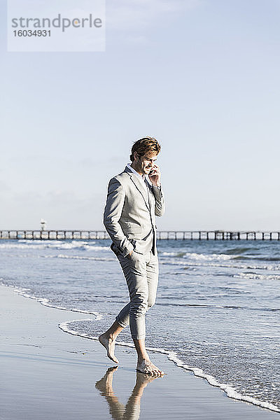Barfüssiger Geschäftsmann spricht mit einem Smartphone am sonnigen Strand des Ozeans,  Los Angeles,  Kalifornien