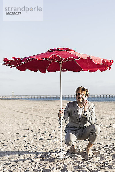 Geschäftsmann spricht mit einem Smartphone unter einem Sonnenschirm am sonnigen Strand,  Los Angeles,  Kalifornien