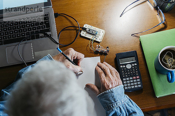 Ausgeschnittenes Bild eines älteren Mannes,  der bei der Reparatur eines Laptops zu Hause ein Diagramm auf Papier erstellt