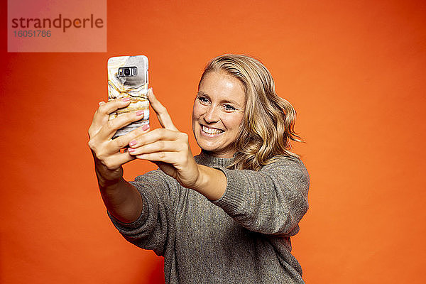 Glückliche blonde Frau nimmt Selfie gegen orange Hintergrund