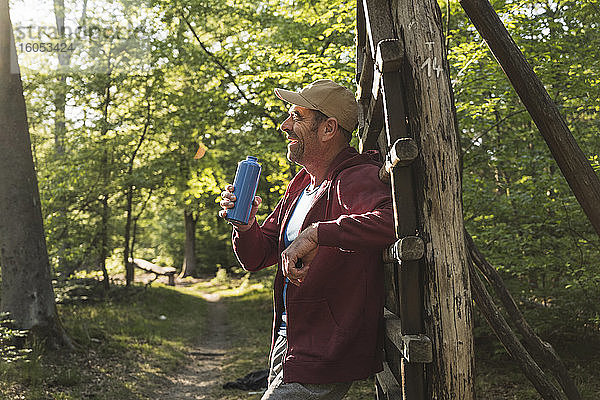 Lächelnder reifer Mann,  der eine Wasserflasche hält,  während er an einer Holzleiter im Park steht