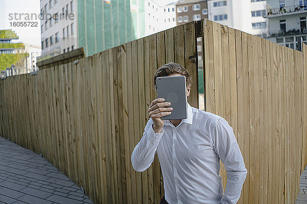 Geschäftsmann bei einem Boarding in der Stadt,  der sein Gesicht mit einem Tablet bedeckt