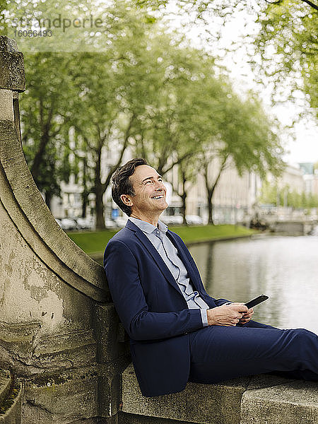 Glücklicher reifer Geschäftsmann mit Smartphone,  der auf einer Brückenbrüstung sitzt
