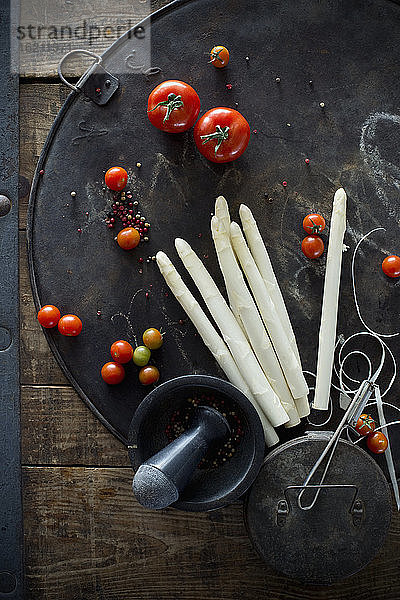 Mörser und Stößel,  Pfefferkörner,  Tomaten und geschälte Spargelstangen auf rustikalem Backblech