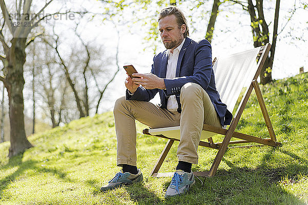 Männlicher Unternehmer,  der sein Smartphone benutzt,  während er auf einem Stuhl im Park sitzt